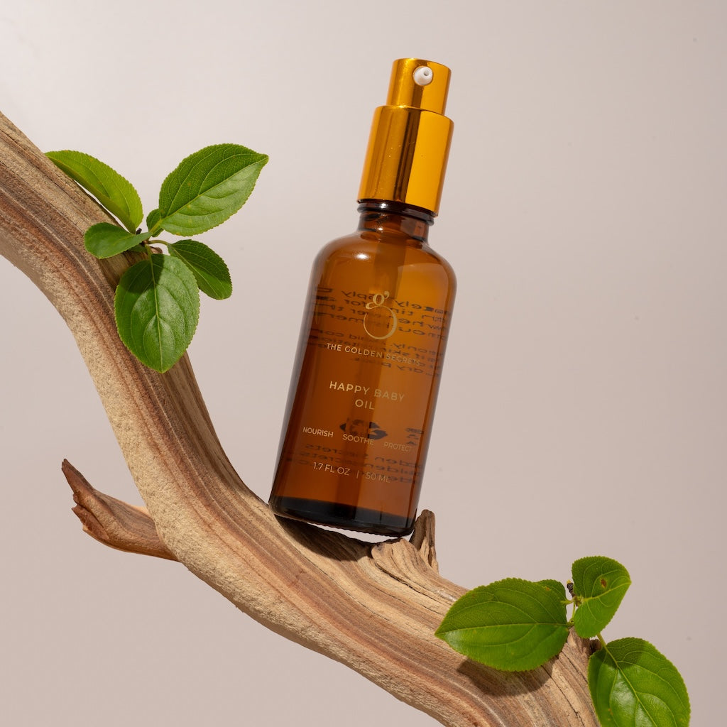 A brown glass bottle with a gold pump labeled Happy Baby Oil rests on a curved branch with green leaves, set against a light beige background.