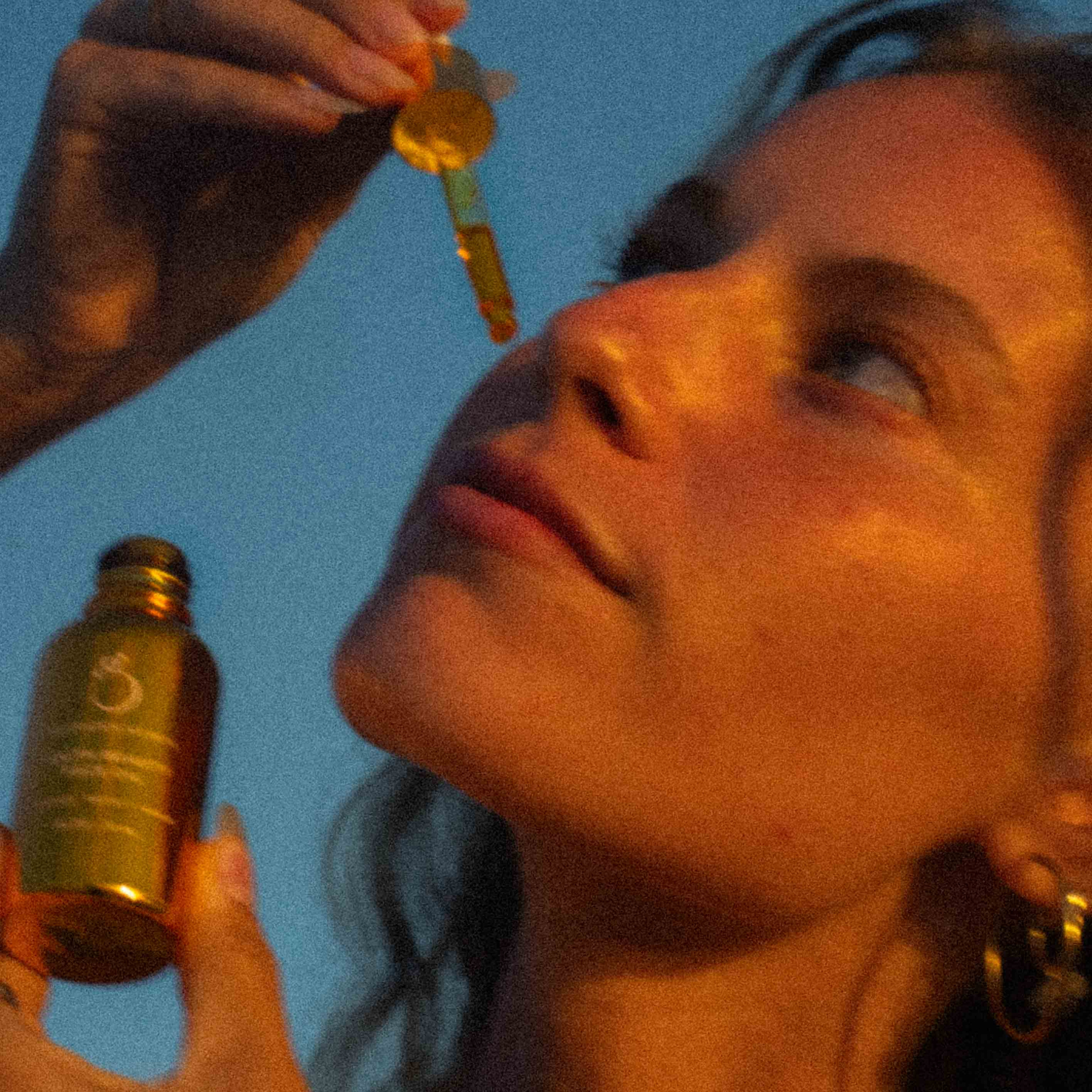 A woman applies a golden facial serum to her face using a dropper, holding a small amber bottle in one hand, with warm sunlight illuminating her skin.