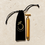 The Gold Pulse Firming Face Wand by THE GOLDEN SECRETS—a 24k gold-plated sonic tool—rests beside a black velvet pouch adorned with the brand’s logo on a cream-colored stone surface, ready to lift and sculpt your skin while supporting lymphatic drainage.