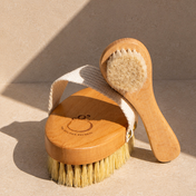The Dry Brush Duo is pictured on a light surface: one brush with a handle and soft bristles, and the other with a fabric strap and firmer bristles for exfoliation and lymphatic support. Sunlight gently highlights both brushes.