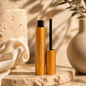 A gold tube of THE GOLDEN SECRETS Long Lash Growth Serum, its applicator wand open, rests on a stone surface accented by soft fabric and neutral décor in the background, all warmed by sunlight—promising fuller lashes with every use.