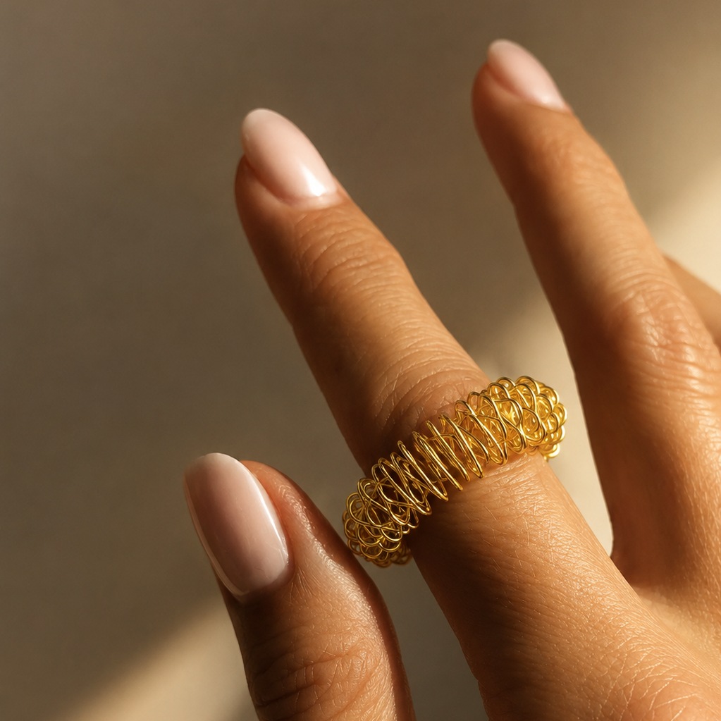 A hand with light pink manicured nails wears THE GOLDEN SECRETS Gold Reflexology Acupressure Massage Ring on the middle finger, set against a softly lit, neutral background—perfect for stimulating finger pressure points while adding a stylish touch.