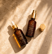 Two amber glass bottles with gold caps, labeled as part of the Sculpt & Glow Duo by THE GOLDEN SECRETS, rest on a textured beige surface bathed in soft sunlight and gentle shadows. These luxurious glow body oils are specially formulated to nourish your skin, enhance its natural radiance, and help diminish the appearance of stretch marks—leaving you luminous all day long.