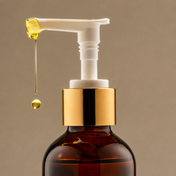 A close-up of THE GOLDEN SECRETS Womb Wisdom Skin-Toning Oil, a brown bottle with a gold and white pump dispenser releasing a drop of yellow, cocoa butter-enriched liquid against a plain beige background—specially formulated to help diminish the appearance of aging for visibly firmer skin.
