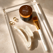 A clear tray holds a gold-lidded facial mask jar, a gold-handled brush, THE GOLDEN SECRETS Goddess Hair Clip in white marble, and a pair of gold hoop earrings on a beige surface—curated for your self-care beauty ritual and glowing skin.