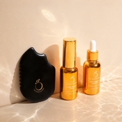 Three skincare products from THE GOLDEN SECRETS' Ultimate Glow Trio Kit are arranged on a beige background: a black Gua Sha tool featuring a gold logo, a shimmering gold spray bottle, and an elegant gold dropper bottle. Light reflections play across the surface, accentuating the Ultimate Glow Trio Kit designed to reveal radiant skin.