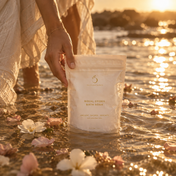A hand holds a THE GOLDEN SECRETS Royal Codes Bath Soak bag in shallow, sunlit water surrounded by flower petals, with a golden sunset and rocks in the background—an atmosphere of tranquility elevated by this mineral-rich bath soak made with pure Zechstein Magnesium Chloride.