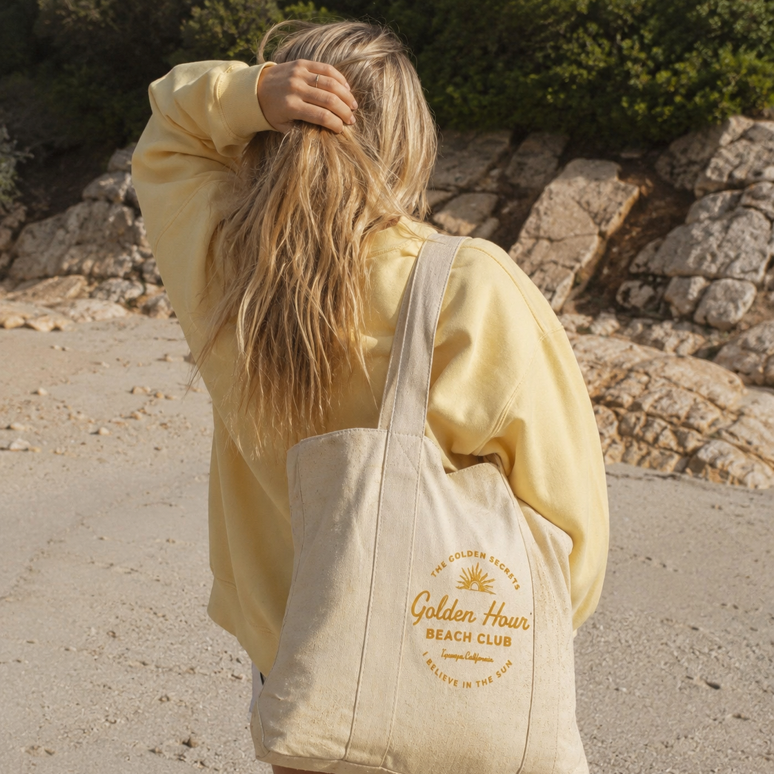 A person with long blond hair, dressed in a yellow sweatshirt, stands on a rocky beach facing away from the camera and holding their hair. They carry THE GOLDEN SECRETS’ Golden Hour Beach Club Tote—a limited-edition natural cotton bag featuring the “Golden Hour Beach Club” design.
