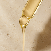 A dropper releasing a stream of THE GOLDEN SECRETS Goddess Shaping Body Oil onto a beige stone surface, formulated to help tighten skin and reduce cellulite.