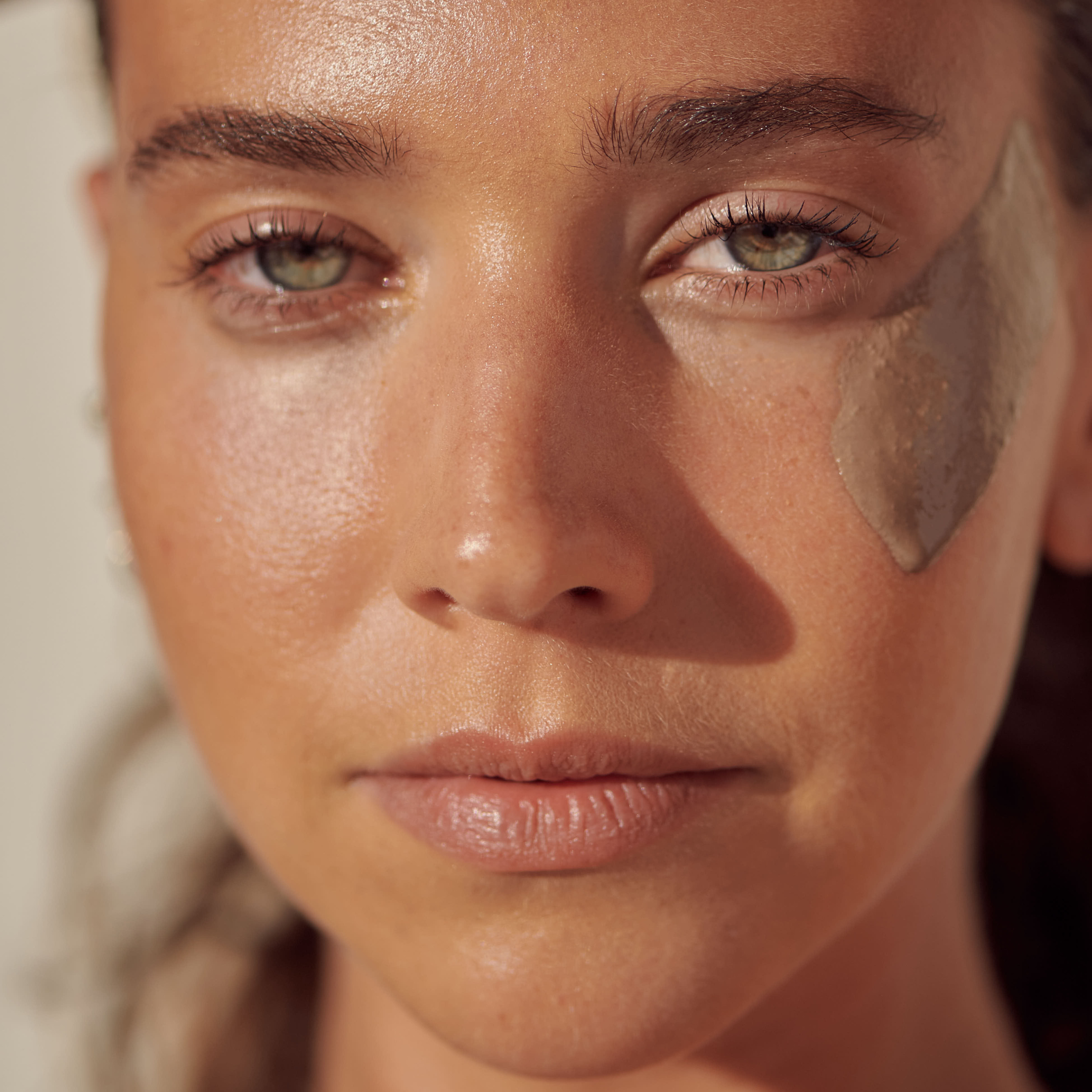 A woman with green eyes and dewy skin wears a patch of THE GOLDEN SECRETS Instant Glow Facial Mask under her right eye, looking directly at the camera in natural light.