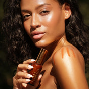 A woman with dark curly hair holds a small amber bottle with a gold cap, while sunlight casts a swatch of orange-tan makeup on her shoulder. The background is a soft, out-of-focus green outdoor setting.