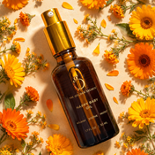 A brown glass bottle of THE GOLDEN SECRETS Happy Baby Oil with a gold spray top rests on a tan surface, surrounded by vibrant marigold petals—perfect to soothe dryness and care for sensitive skin.