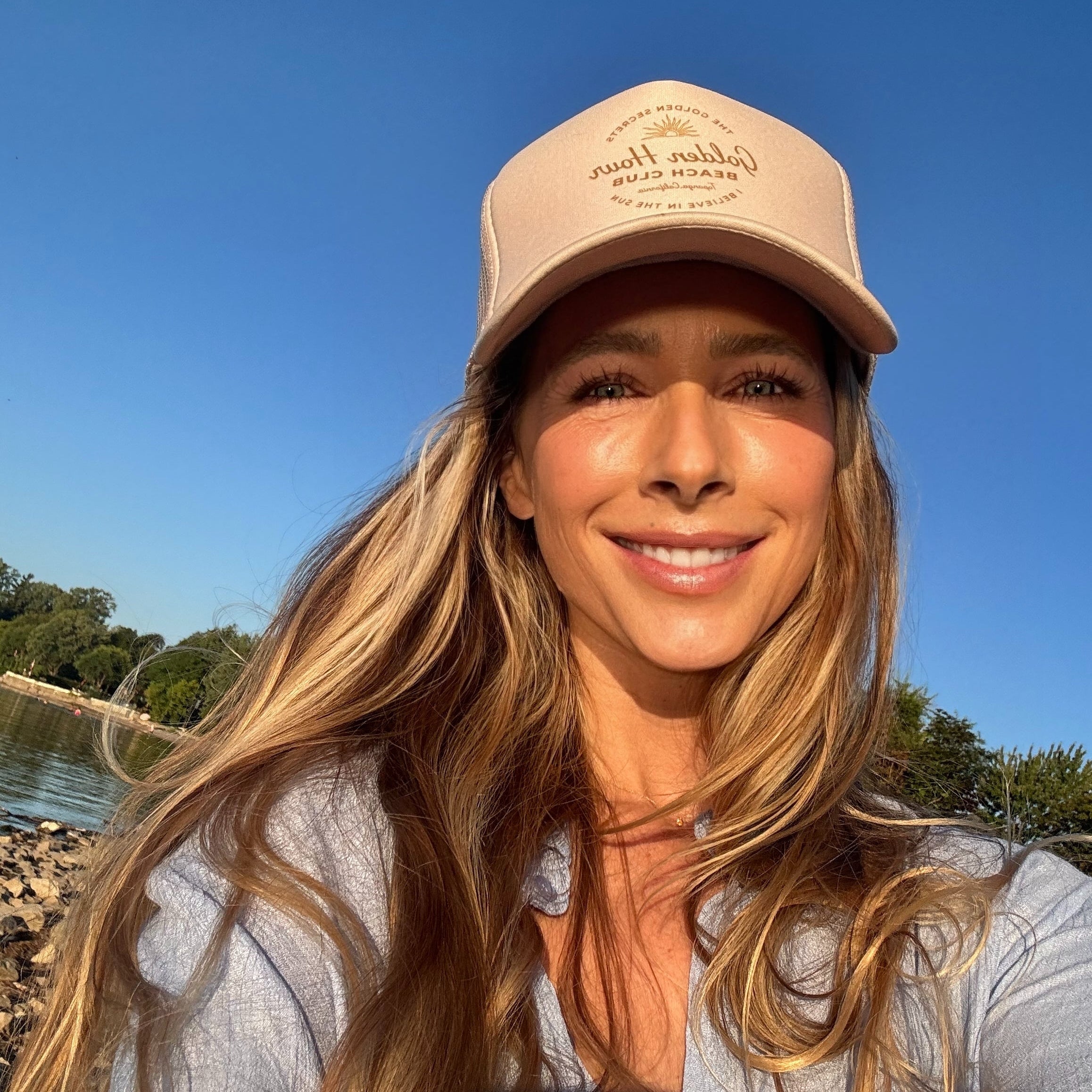 A woman with long blonde hair smiles outdoors by a lake, basking in the warm glow of golden hour and surrounded by nature, wearing a beige “The Golden Secrets Trucker Hat” from THE GOLDEN SECRETS and a light blue shirt against a backdrop of trees and clear blue sky.