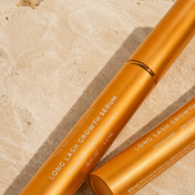 Two gold tubes labeled “Long Lash Growth Serum” by THE GOLDEN SECRETS rest on a textured beige surface, their packaging displaying product details and instructions—presenting this serum as an effective eyelash enhancer for achieving fuller lashes.