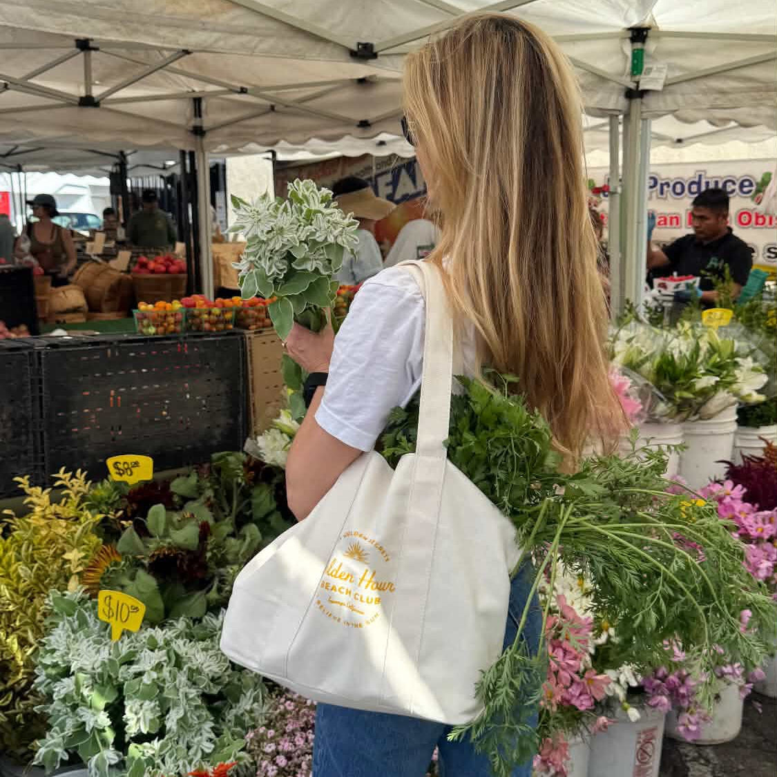 A woman with long blonde hair holds leafy greens at a farmers market. She carries THE GOLDEN SECRETS Golden Hour Beach Club Tote over her shoulder and is surrounded by flowers and produce stands under white tents.