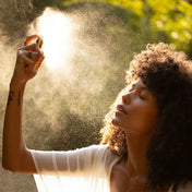 A woman with curly hair sprays a fine mist from a small bottle into the air, with sunlight and green foliage softly blurred in the background.