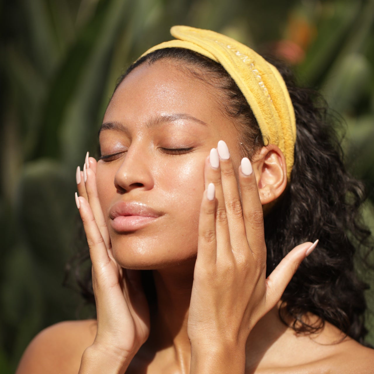 A woman wearing a yellow headband gently touches her radiant face with both hands, eyes closed, standing outdoors against a blurred green background—her glowing complexion hinting at the refreshing effects of THE GOLDEN SECRETS Magic Brightening Powder 3-in-1.