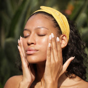 A woman wearing a yellow headband gently touches her radiant face with both hands, eyes closed, standing outdoors against a blurred green background—her glowing complexion hinting at the refreshing effects of THE GOLDEN SECRETS Magic Brightening Powder 3-in-1.