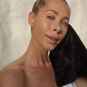 A woman with blue eyes and light skin holds a Glowing Facial Cloth by THE GOLDEN SECRETS to her cheek, posing against a neutral background. Her hair is pulled back, and she is wearing minimal makeup and a strapless top.