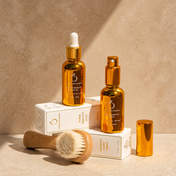 The Lymph & Lift Kit, featuring two gold bottles and a nourishing face oil for deep hydration, is displayed on white boxes with a wooden facial dry brush and gold cap nearby, all set against a beige background.