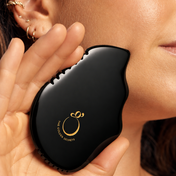 A close-up lifestyle shot of a woman holding a black, teardrop-shaped facial massage tool against her cheek. The tool features a gold logo that reads 'THE GLOWING SECRETS' and is held by her hand.