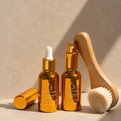 The Lymph & Lift Kit, featuring a golden face oil bottle with a dropper and a spray bottle, is displayed on a beige surface next to a wooden dry brushing brush in bright, softly shadowed lighting.