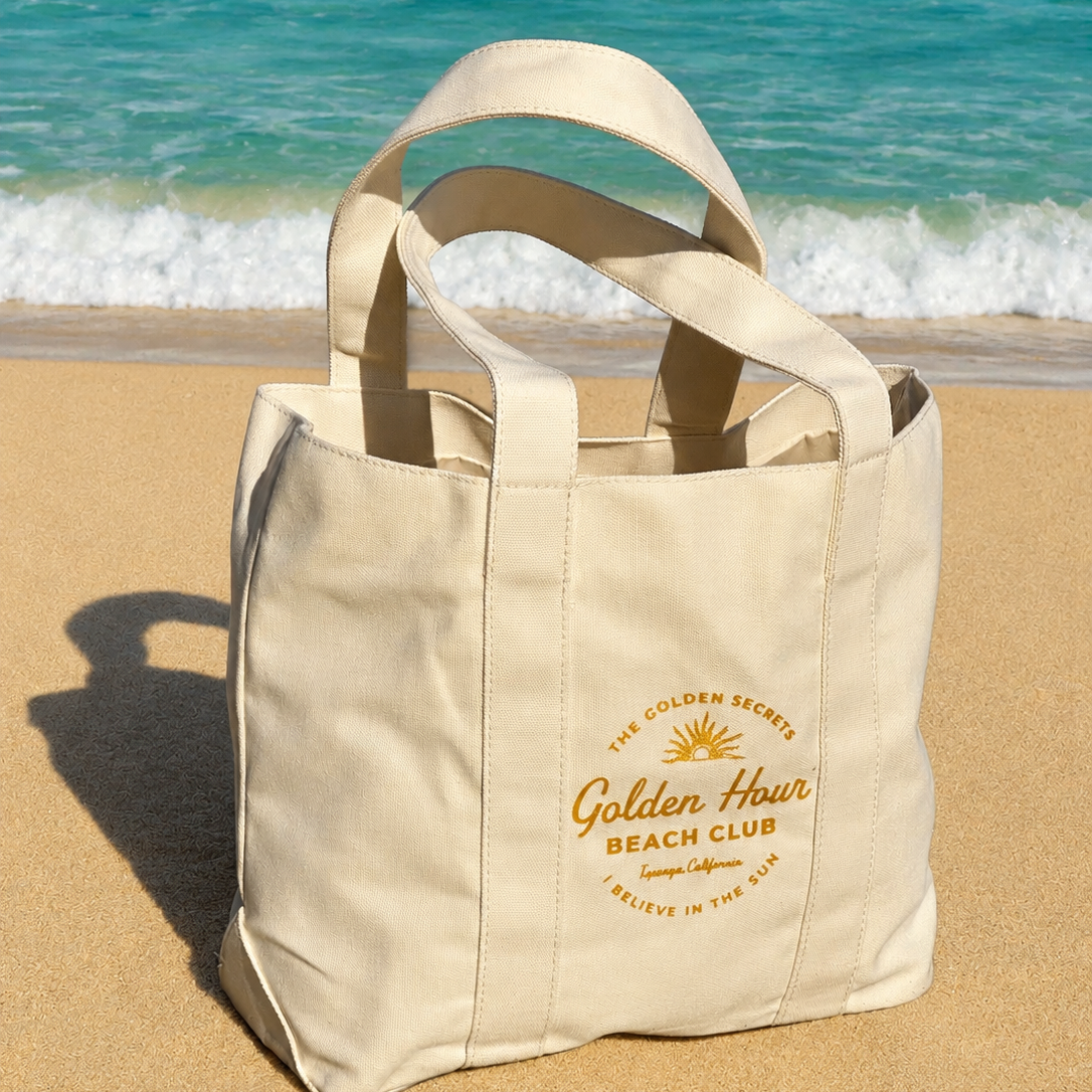Beige tote bag with 'Golden Hour Beach Club' text on a sandy beach with ocean waves in the background