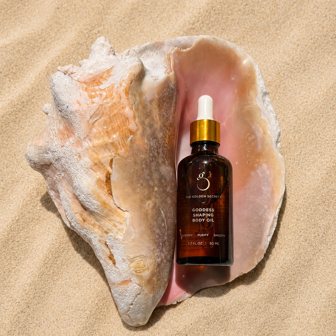 A brown glass dropper bottle of THE GOLDEN SECRETS Goddess Shaping Body Oil rests inside a large seashell on a sandy beach, offering to reduce cellulite and tighten skin for a smoother, more radiant appearance.