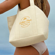 A woman wearing a cream canvas tote bag over her shoulder, photographed outdoors against a soft blue sky and ocean background. The tote bag has a gold printed logo reading 'Golden Hour Beach Club' with 'San Diego, California' below it.
