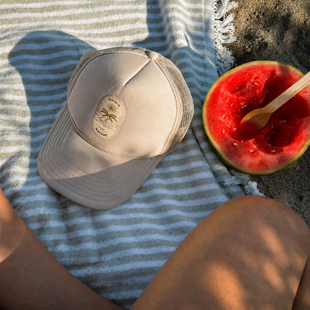A THE GOLDEN SECRETS – The Golden Secrets Trucker Hat in beige rests on a striped towel beside a half-eaten watermelon and spoon atop sandy ground. Bathed in golden hour sunlight, the scene captures part of a person’s legs for a serene, nature-inspired moment.
