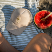 A THE GOLDEN SECRETS – The Golden Secrets Trucker Hat in beige rests on a striped towel beside a half-eaten watermelon and spoon atop sandy ground. Bathed in golden hour sunlight, the scene captures part of a person’s legs for a serene, nature-inspired moment.