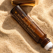 A roll-on essential oil perfume bottle and its cap from 'The Golden Secrets Signature Blend' resting on textured sand.
