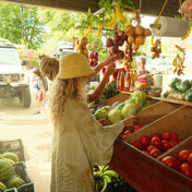 Two shoppers browse fresh produce at a bustling outdoor market, with one donning THE GOLDEN SECRETS Golden Sun Bucket Hat as they pick out cabbages and tomatoes beneath hanging bananas, onions, and garlic illuminated by sunbeams.