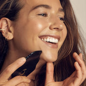 A close-up lifestyle shot of a woman smiling while using a black facial massage device with a gold logo on her cheek.