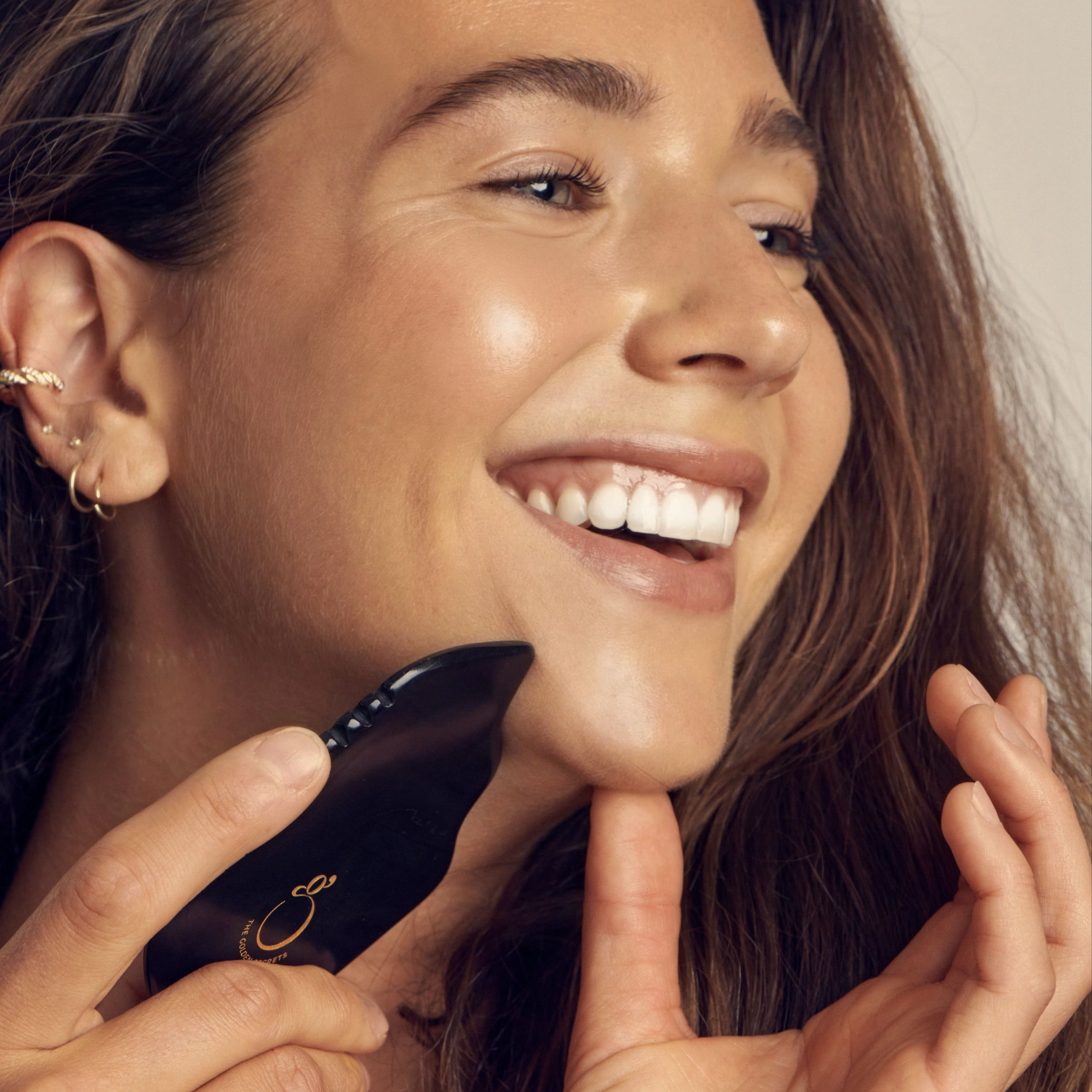 A close-up lifestyle shot of a woman smiling while using a black facial massage device with a gold logo on her cheek.
