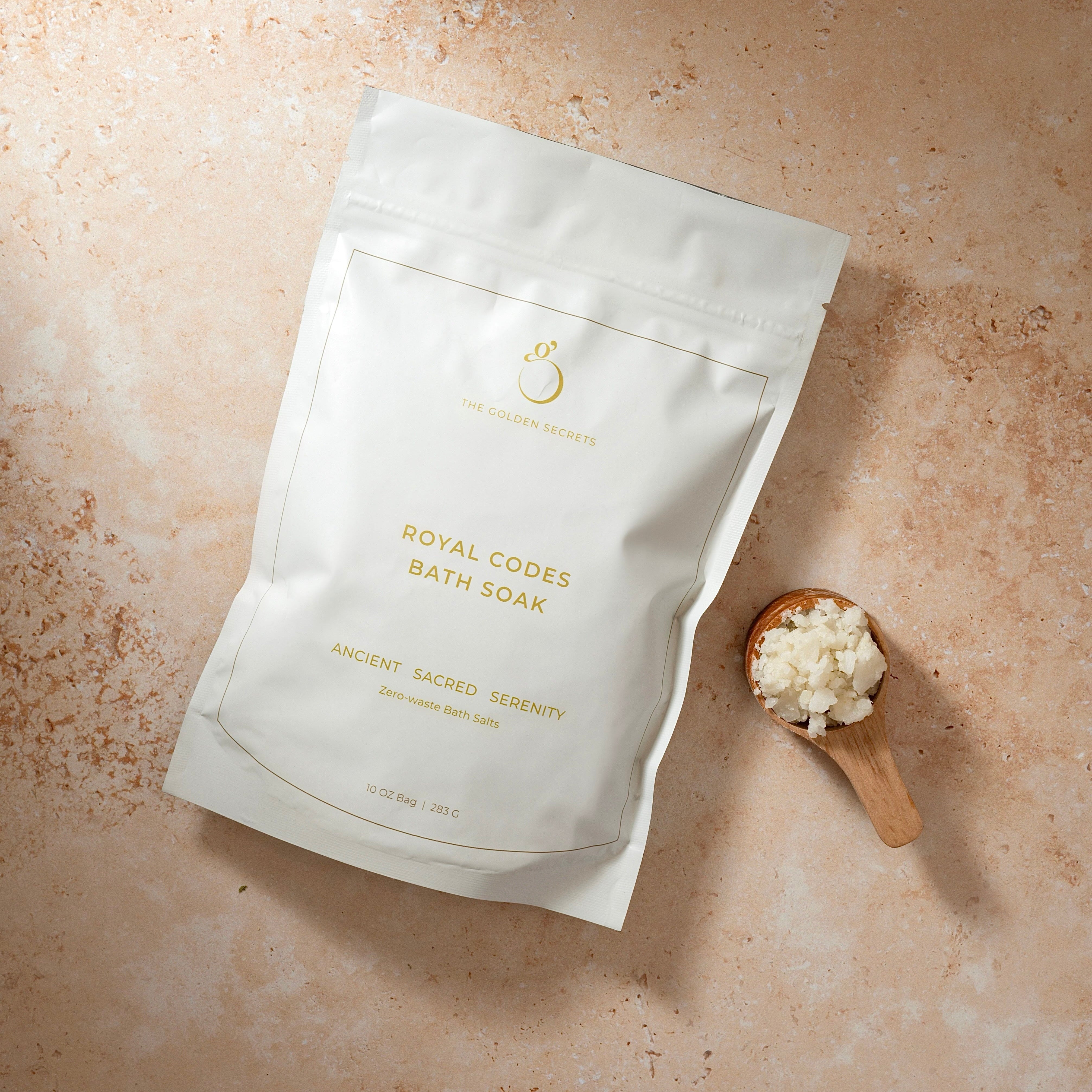 A white pouch labeled Royal Codes Bath Soak by THE GOLDEN SECRETS rests on a beige surface beside a wooden scoop filled with white bath salts. The minimalist gold text and branding on the pouch reflect the premium MSM bath soak blend inside, crafted to elevate your bathing ritual.