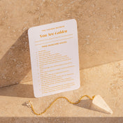 A white card titled 'You Are Golden' from 'The Crown Secrets' lies on a beige stone surface next to a gold chain with a white crystal pointer.