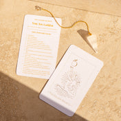 A white tarot card set with gold foil printing, including 'The Golden Secret' and 'You Are Golden', displayed on a textured beige stone surface with a gold chain and white crystal pendant.