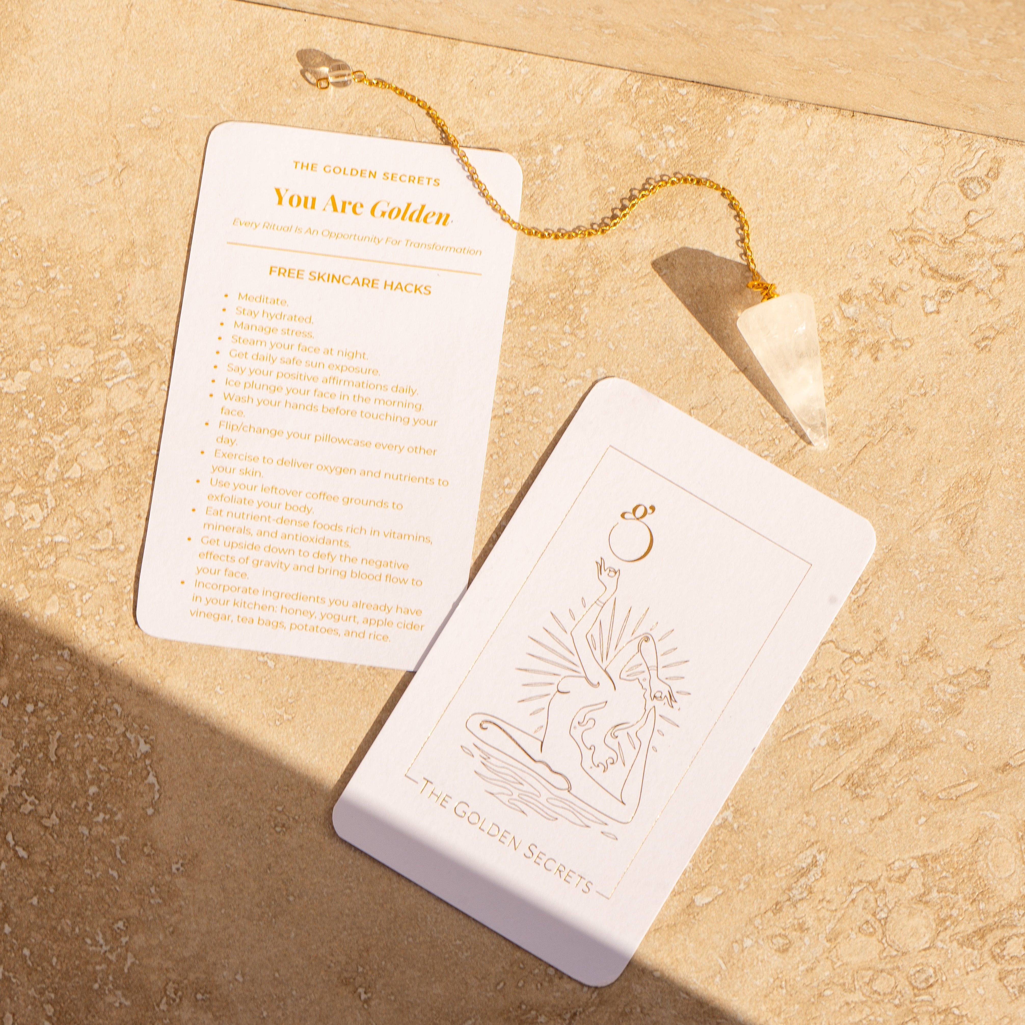A white tarot card set with gold foil printing, including 'The Golden Secret' and 'You Are Golden', displayed on a textured beige stone surface with a gold chain and white crystal pendant.