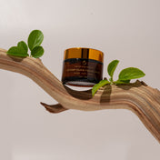 A jar of THE GOLDEN SECRETS Instant Glow Facial Mask, topped with a gold lid, sits elegantly on a curved wooden branch adorned with green leaves, set against a plain beige background.