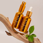 The Dynamic Duo by THE GOLDEN SECRETS — featuring a hydrating mist in a golden spray bottle and a nourishing face oil with a dropper — rests atop a curved wooden piece with a sprig of fresh greenery, set against an understated beige backdrop.