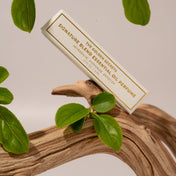 A rectangular box of THE GOLDEN SECRETS Signature Blend Essential Oil is displayed on a twisted piece of wood, surrounded by lush green leaves against a neutral backdrop, inviting you to discover its unique scent and alluring fragrance.