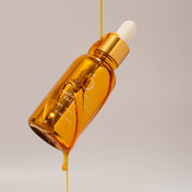 A golden dropper bottle of THE GOLDEN SECRETS Youth Beauty Face Oil, capped in white, tilts as a stream of luxurious golden oil pours gracefully against a plain beige background.