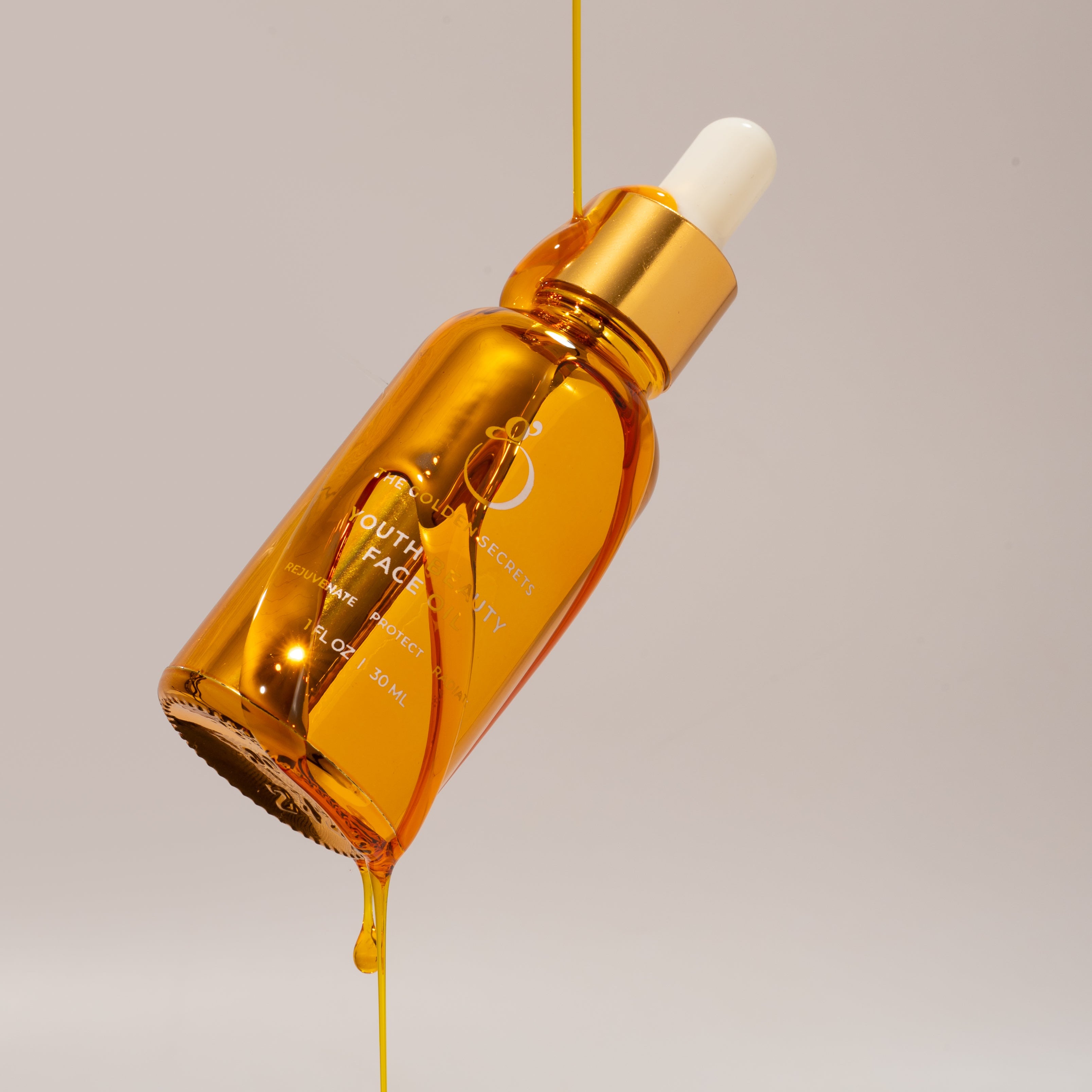 A golden dropper bottle of THE GOLDEN SECRETS Youth Beauty Face Oil, capped in white, tilts as a stream of luxurious golden oil pours gracefully against a plain beige background.