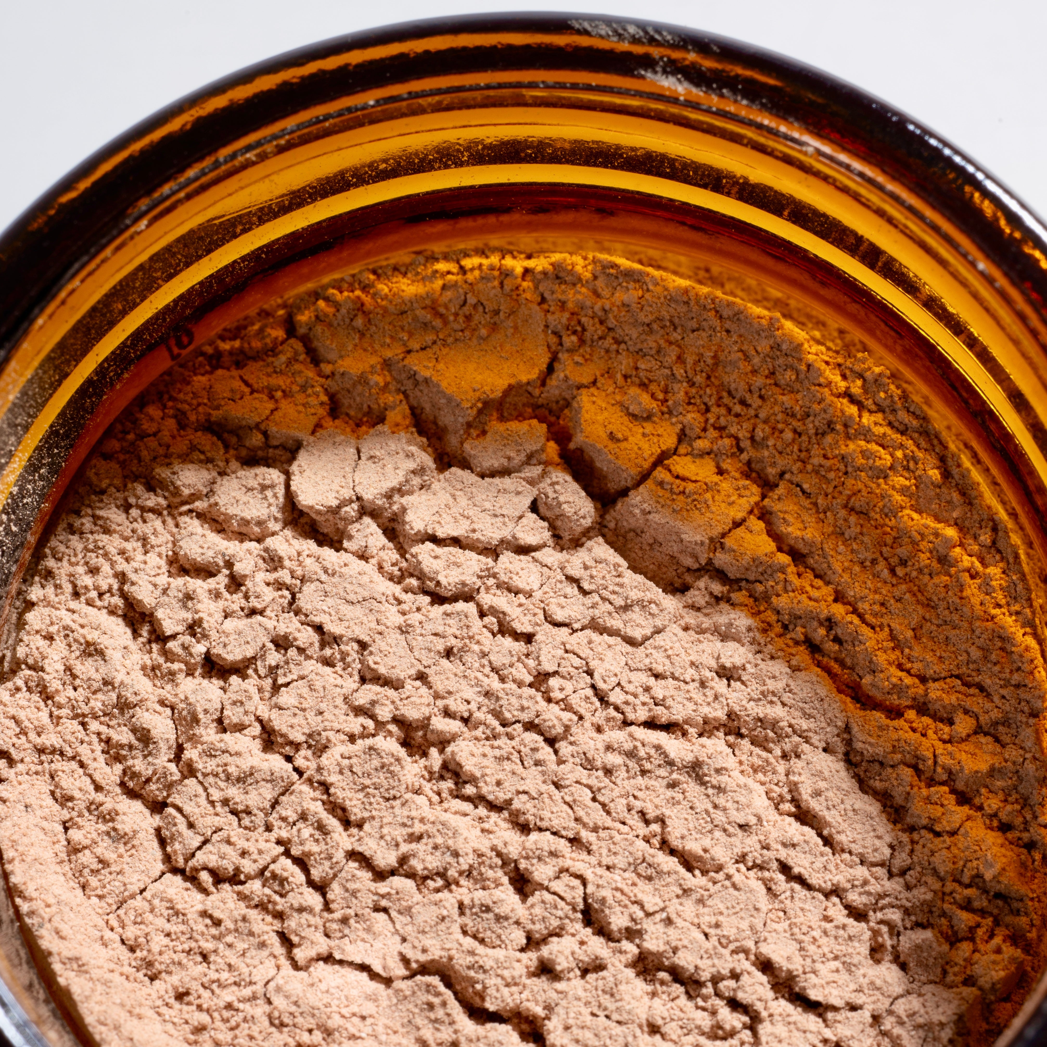 Close-up of a light tan cosmetic powder inside an open amber glass jar, showing the textured surface of the cream or powder against a white background.