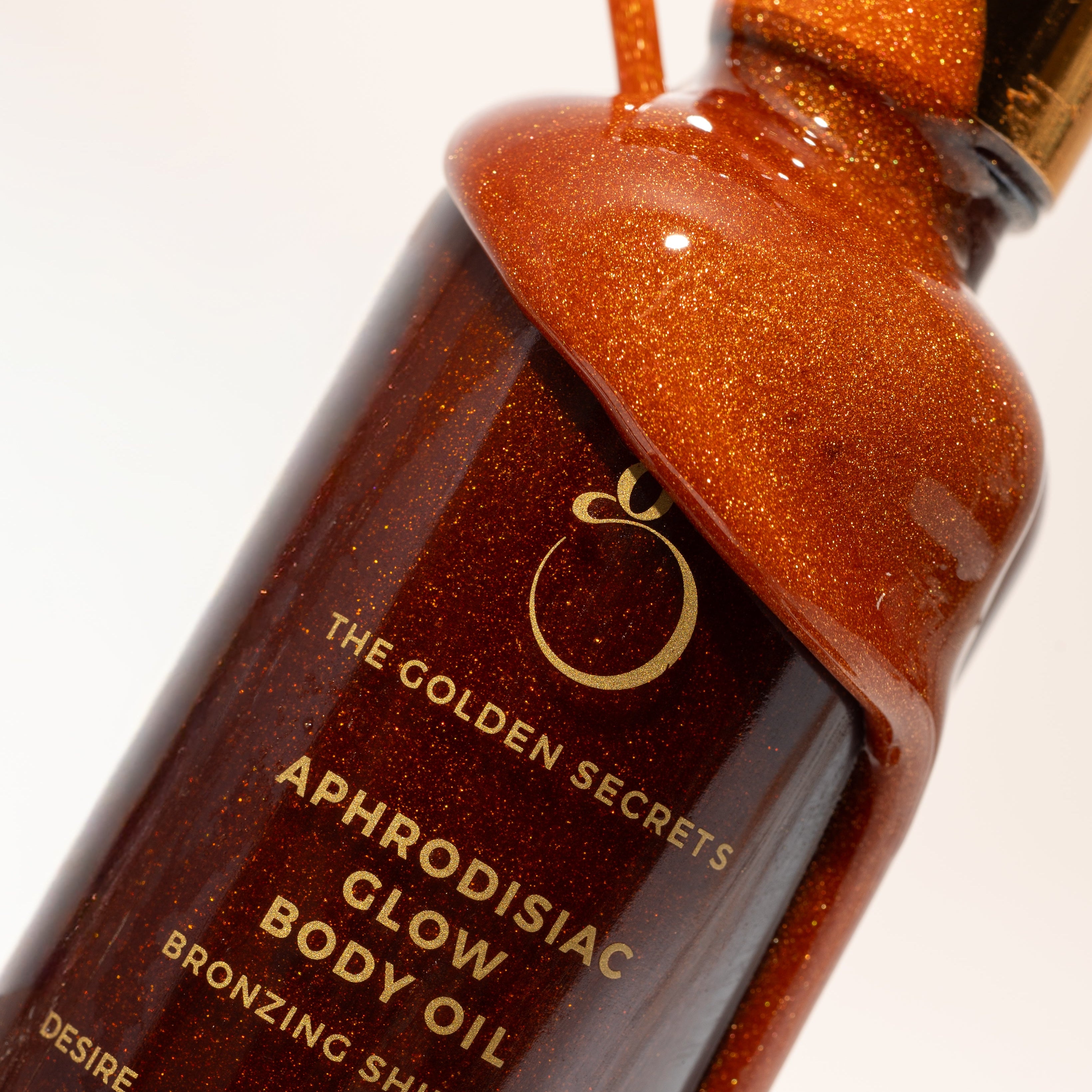 A close-up of a bottle labeled Aphrodisiac Glow Oil from THE GOLDEN SECRETS, with glimmering golden oil spilling over the top. The packaging showcases gold lettering and a radiant, bronzed sheen that hints at the sun-kissed luminosity inside. Infused with jasmine sambac, this natural bronzing oil promises glowing, luminous skin and an irresistible scent.