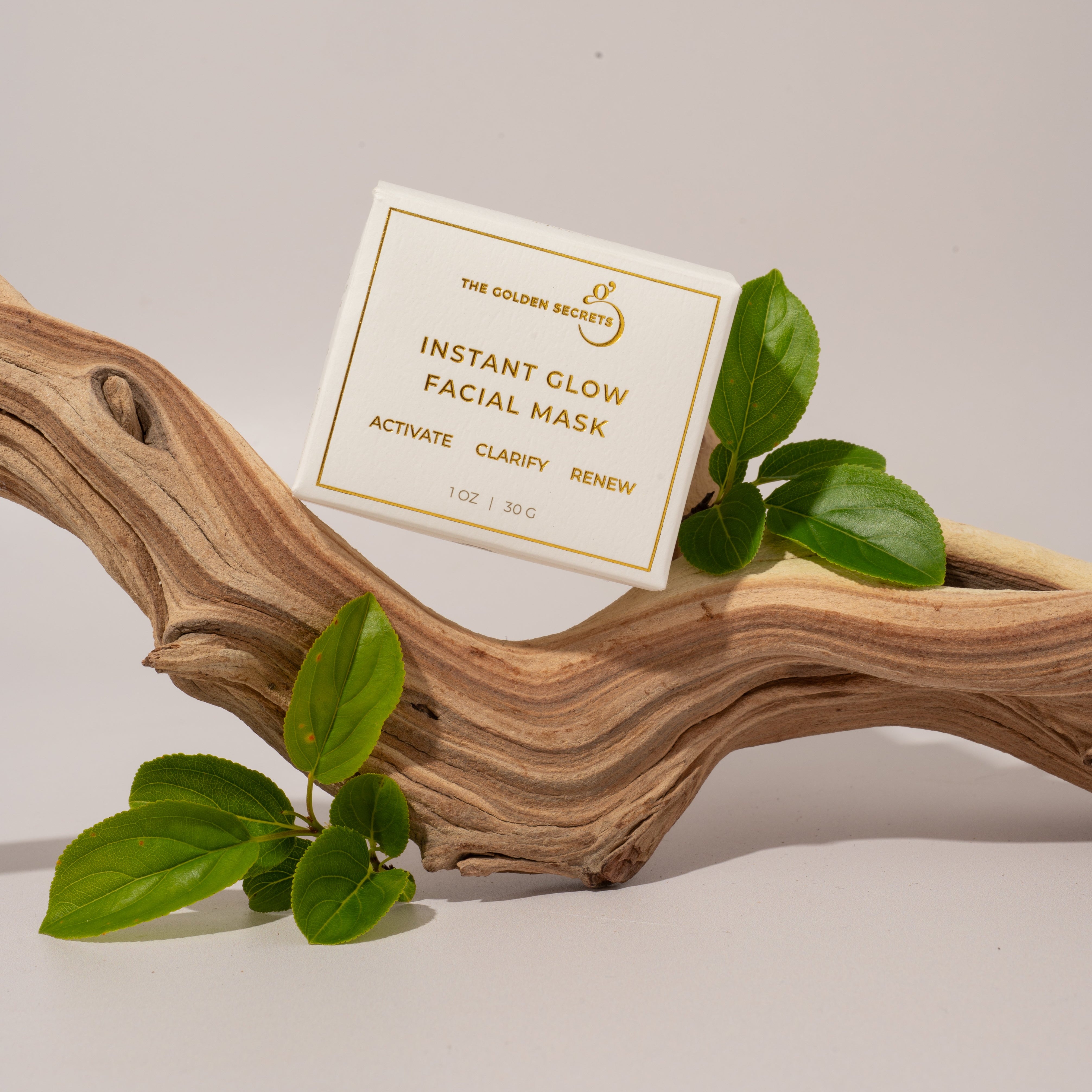 A box of THE GOLDEN SECRETS Instant Glow Facial Mask, an antioxidant-rich treatment perfect for aging skin, sits atop a curved piece of driftwood adorned with green leaves against a neutral backdrop.