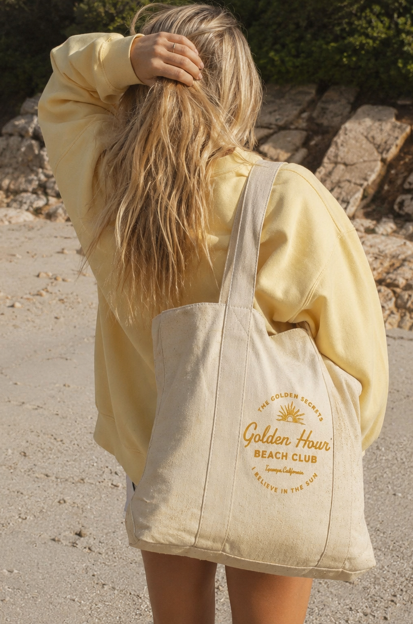 A person with long blonde hair wearing a yellow sweatshirt stands on a sandy beach, holding a beige tote bag that reads Golden Hour Beach Club in yellow text. Rocks and greenery appear in the background.