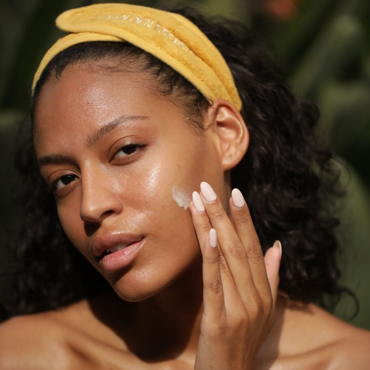 A woman with curly dark hair wearing a yellow headband applies white skincare cream to her cheek, set against a dark green tropical background.