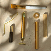 A flat-lay arrangement of The Golden Secrets skincare and beauty tools on a textured beige surface with dappled sunlight and shadows. The set includes a dual-ended brush with a white handle and gold ferrule, a gold facial roller tool, a gold tube, a black-and-gold serum bottle, a gold chain with a small heart pendant, a gold scrunchie, and a wooden stick.